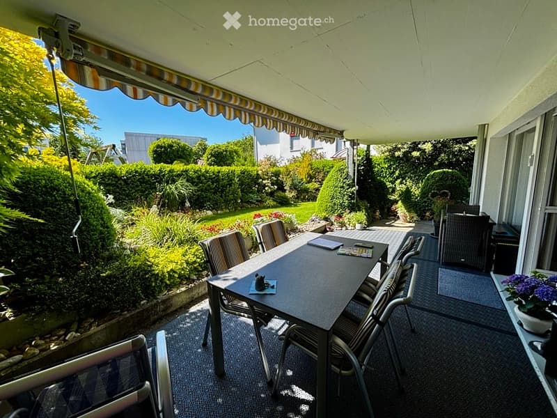A covered balcony with a dining table and chairs, overlooking a well-maintained garden with various plants and trees.