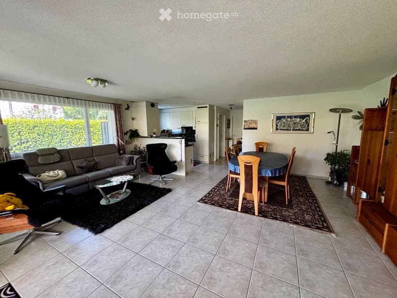 Open-plan living and dining area with a grey sofa, coffee table, dining table with chairs, and a modern kitchen area in the background.