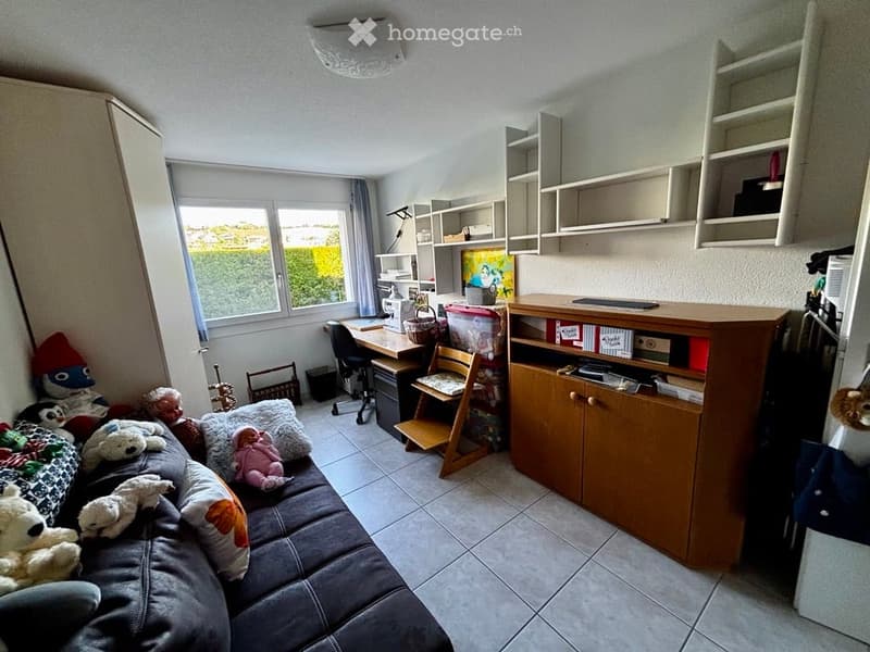 A cozy, multi-functional room with a grey sectional sofa adorned with pillows and stuffed animals, a wooden desk with a chair, a cabinet with shelves, and built-in storage units. The room features large windows with a view of greenery outside.