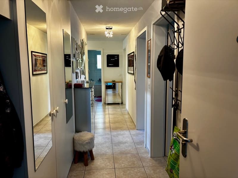 A well-lit hallway with tiled flooring, white walls, and several doors leading to different rooms. There are framed pictures on the walls, a small wooden stool with a fur cushion, and a coat rack with jackets hanging on it.