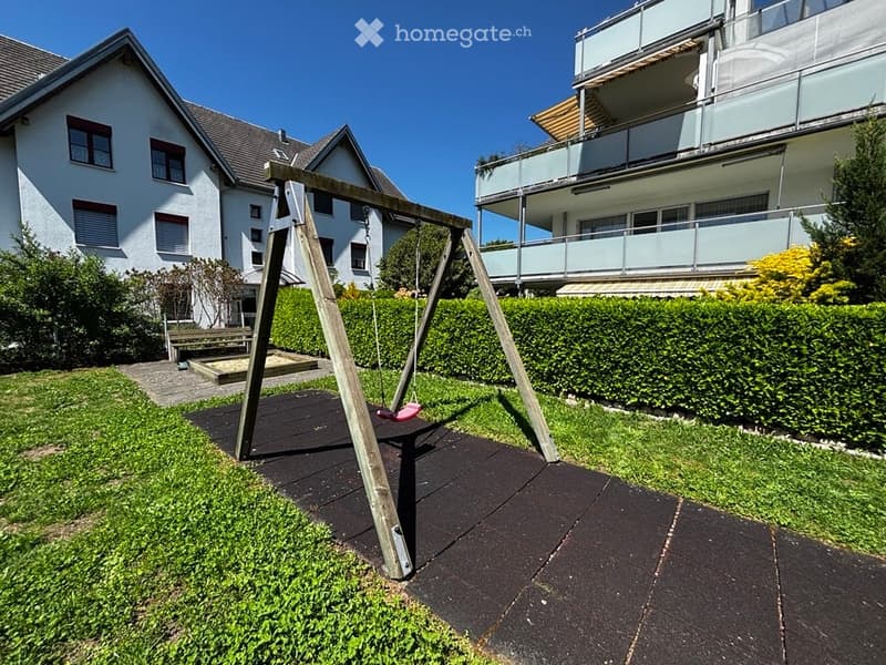 A small playground area with a swing set and a bench, surrounded by neatly trimmed hedges and grass, with residential buildings in the background.