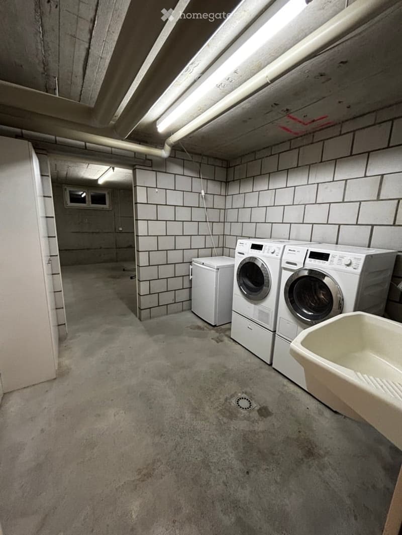 Underground laundry room with concrete floor, tiled walls, fluorescent lighting, and a small window. Contains a washing machine, dryer, and a sink.