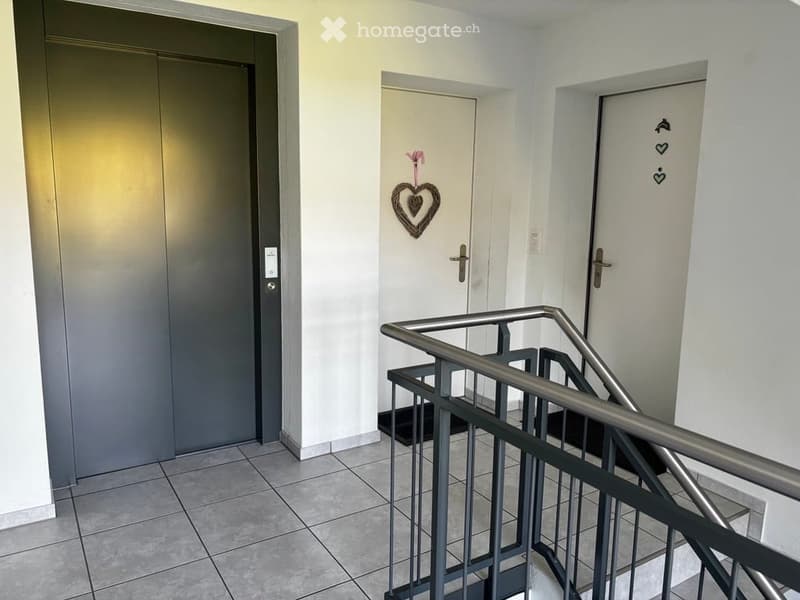 A clean, modern hallway with tiled flooring, white walls, and three doors. A heart-shaped decorative item hangs on one door, and a metal railing leads to an upper level.
