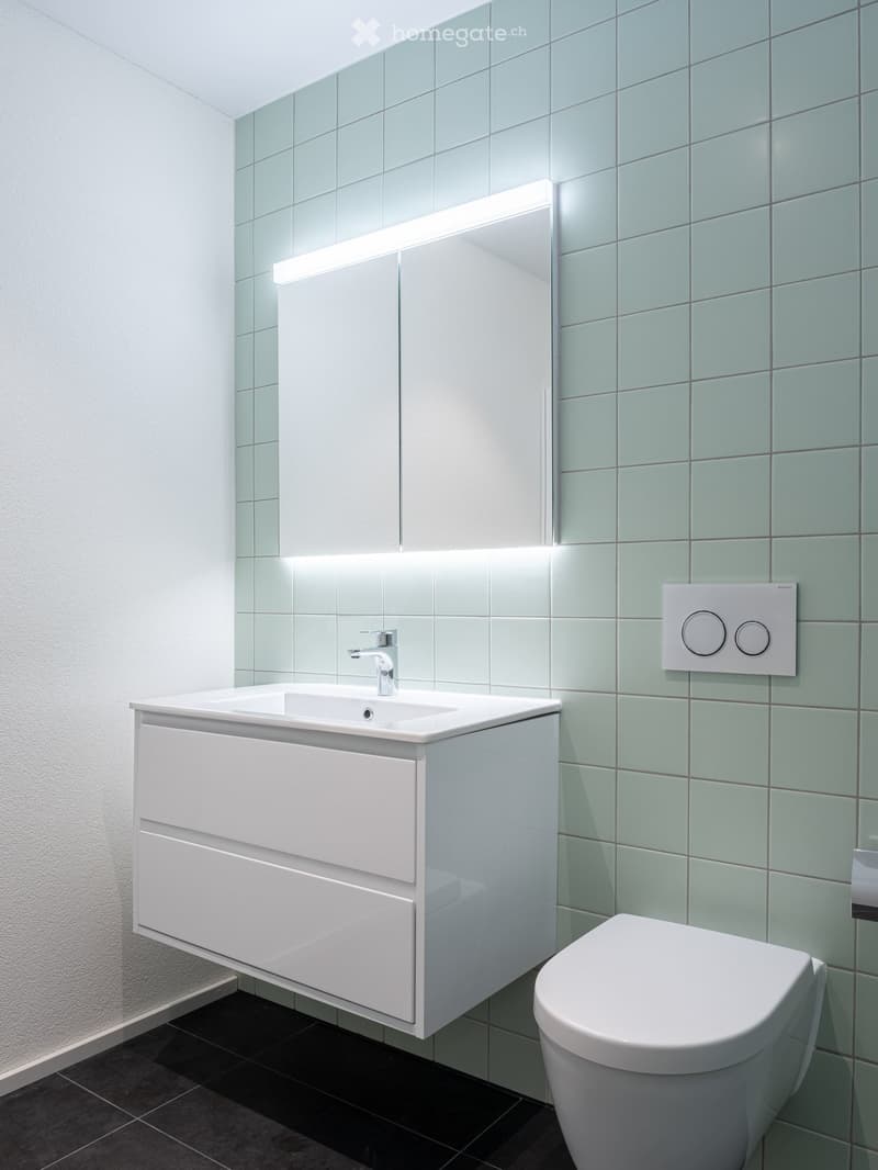 Modern bathroom with white tiles, a white sink with a chrome faucet, a large mirror with integrated lighting, a white toilet, and a white cabinet with drawers.