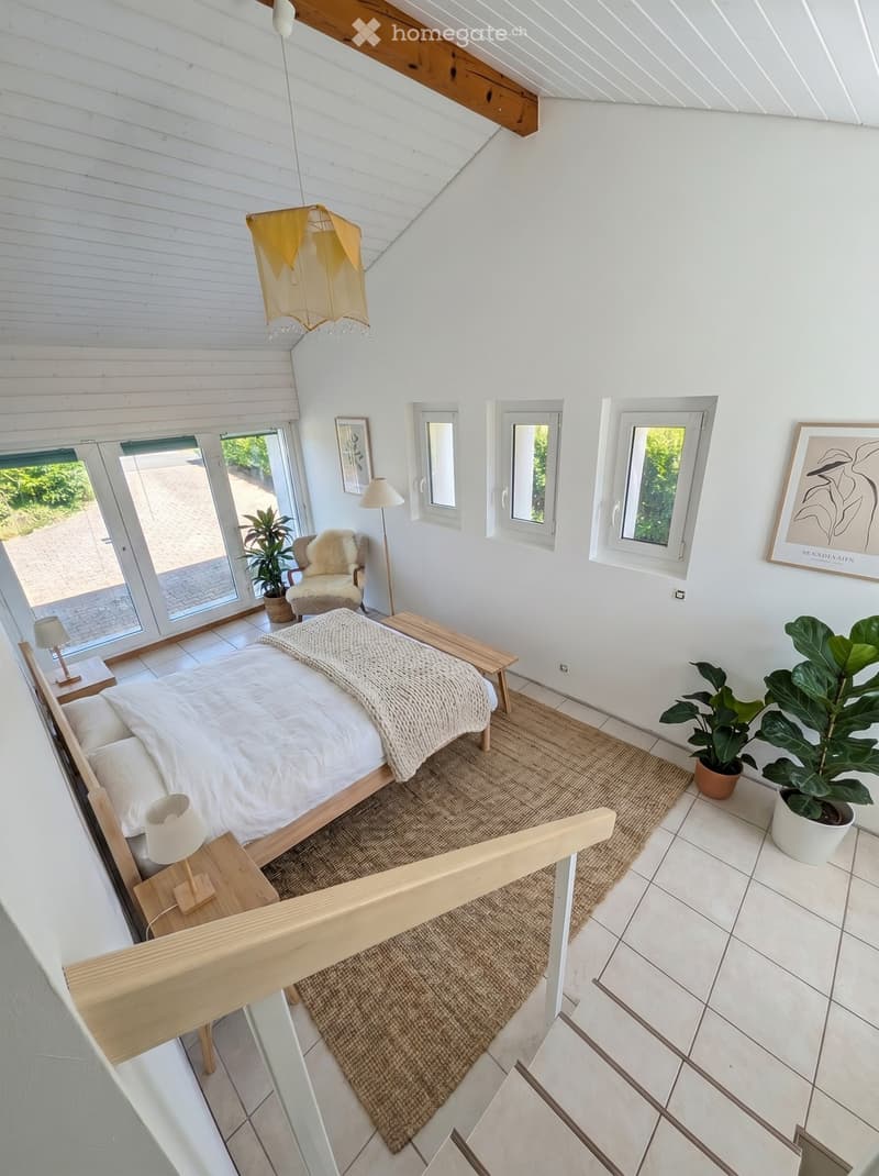Bright, modern bedroom with white walls, wooden bed frame, nightstands, lamps, a cozy chair, a bench, a large potted plant, and artwork on the walls.