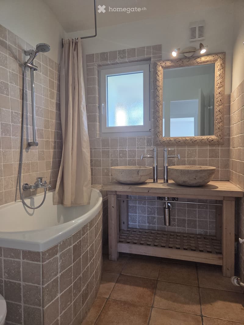 Bathroom with a bathtub, shower head, and a large mirror above a window. The room features tiled walls and floor, with a shower curtain partially drawn.