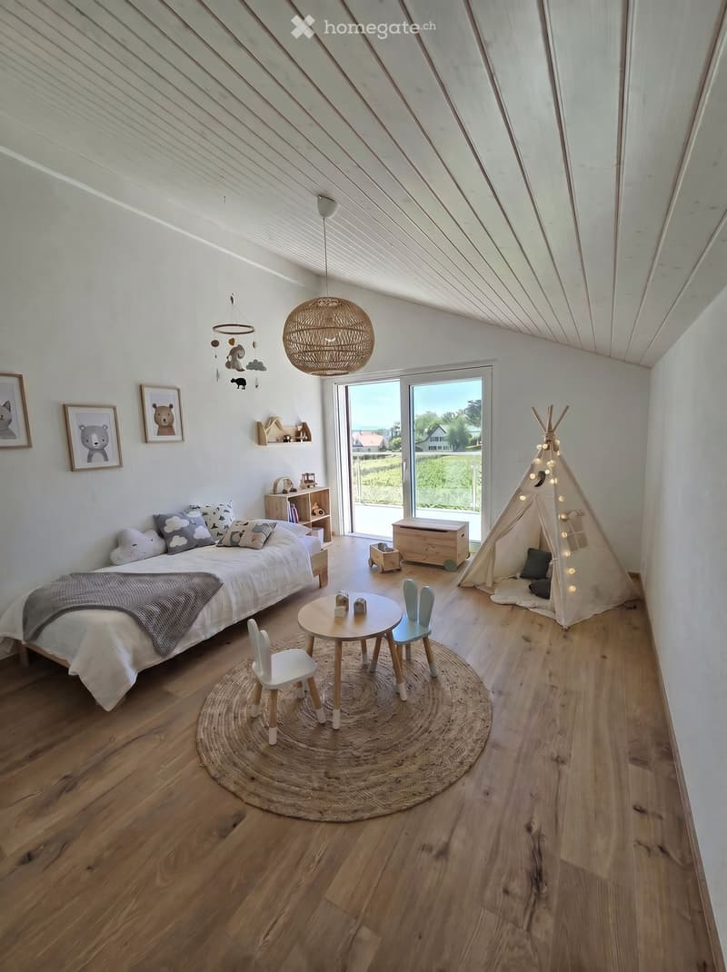 Bright, airy bedroom with wooden floors, a large bed, a small round table with chairs, a teepee tent, and a hanging wicker lamp. Large windows offer a view of the outdoors.
