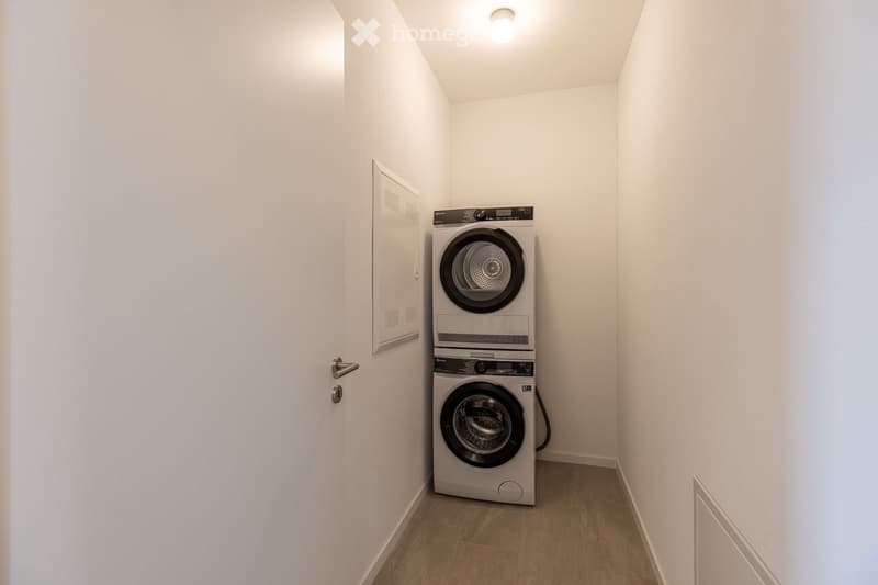 A small, empty room with white walls and wooden flooring. It contains a stack of two washing machines and a light on the ceiling.