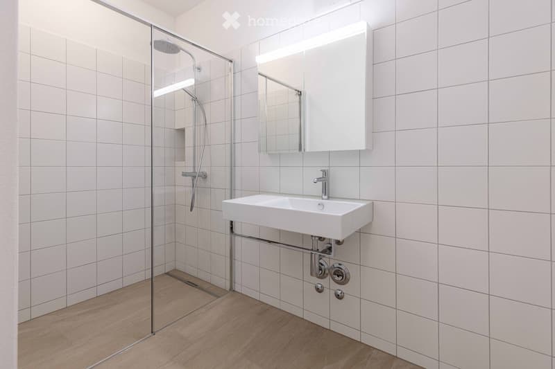 Modern bathroom with white tiles, glass shower enclosure, white sink, and a mirror with a built-in light.