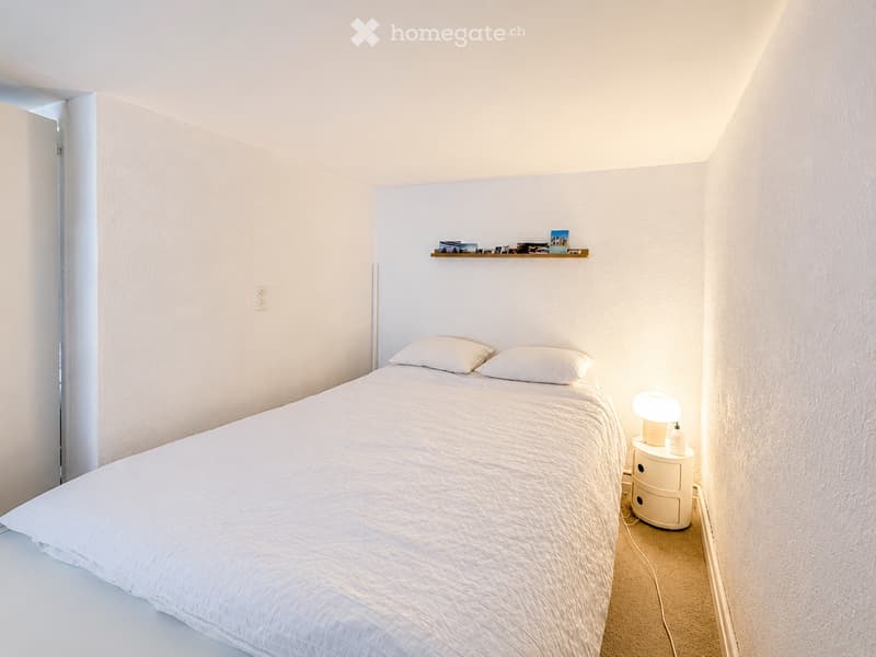 Minimalist bedroom with a white bed, nightstand, lamp, and a small shelf with photos and decorative items.