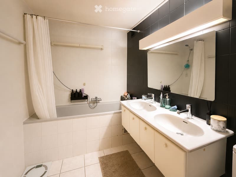 Modern bathroom with a bathtub, double sink, large mirror, and a shower area with a curtain. Includes a bathroom scale and a rug on the floor.