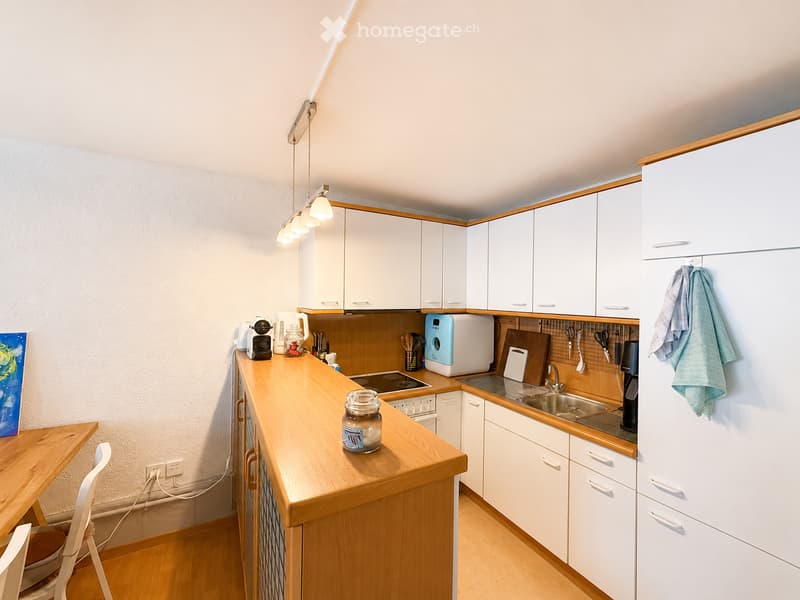 Modern kitchen with white cabinets, wooden countertops, and stainless steel appliances. Features a sink, microwave, coffee maker, and a small dining area with a chair and table.