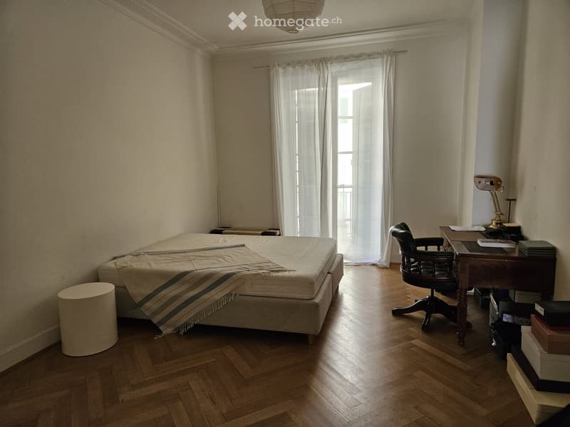 Bedroom with wooden floor, large window with white curtains, bed with striped blanket, desk with chair, and lamp.