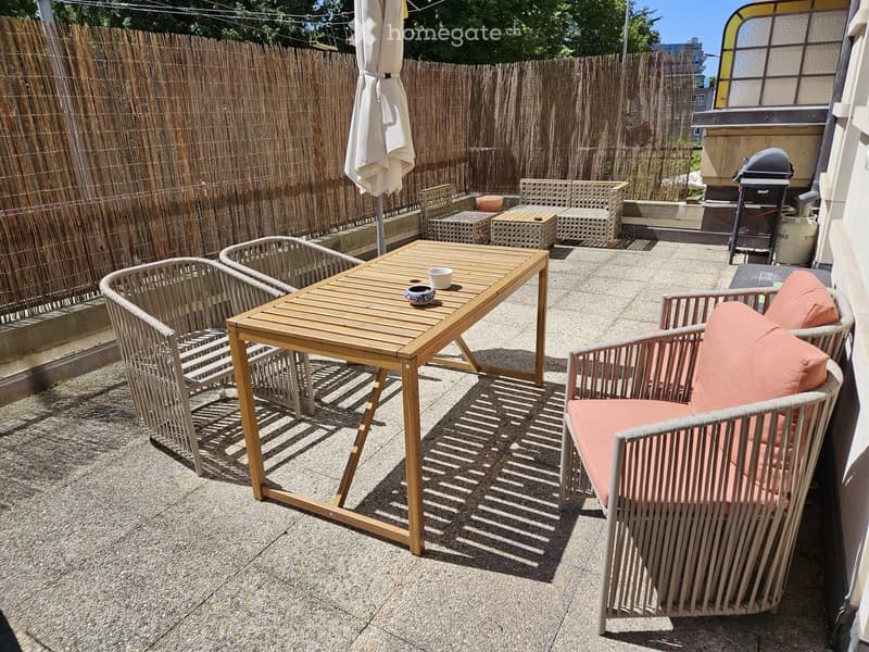 A small outdoor patio with a wooden table, wicker chairs, and an umbrella, enclosed by a bamboo fence.