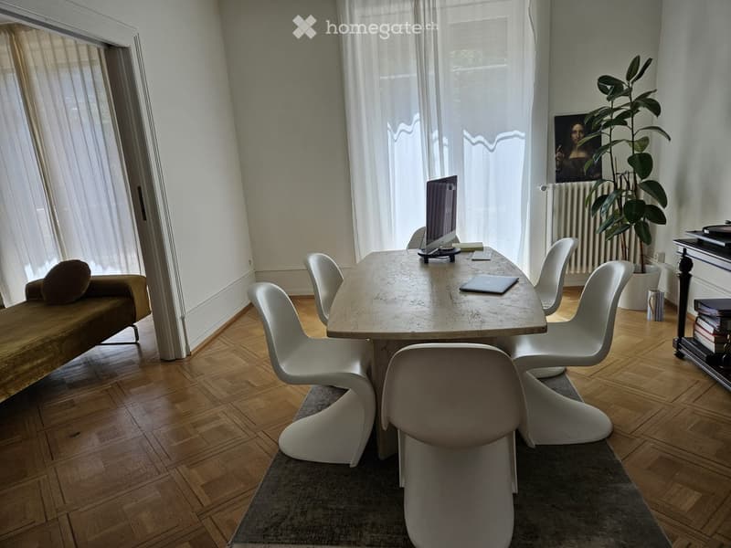 Bright room with wooden flooring, large windows with sheer curtains, a sofa, a dining table with chairs, a laptop, a potted plant, and a picture frame on the wall.