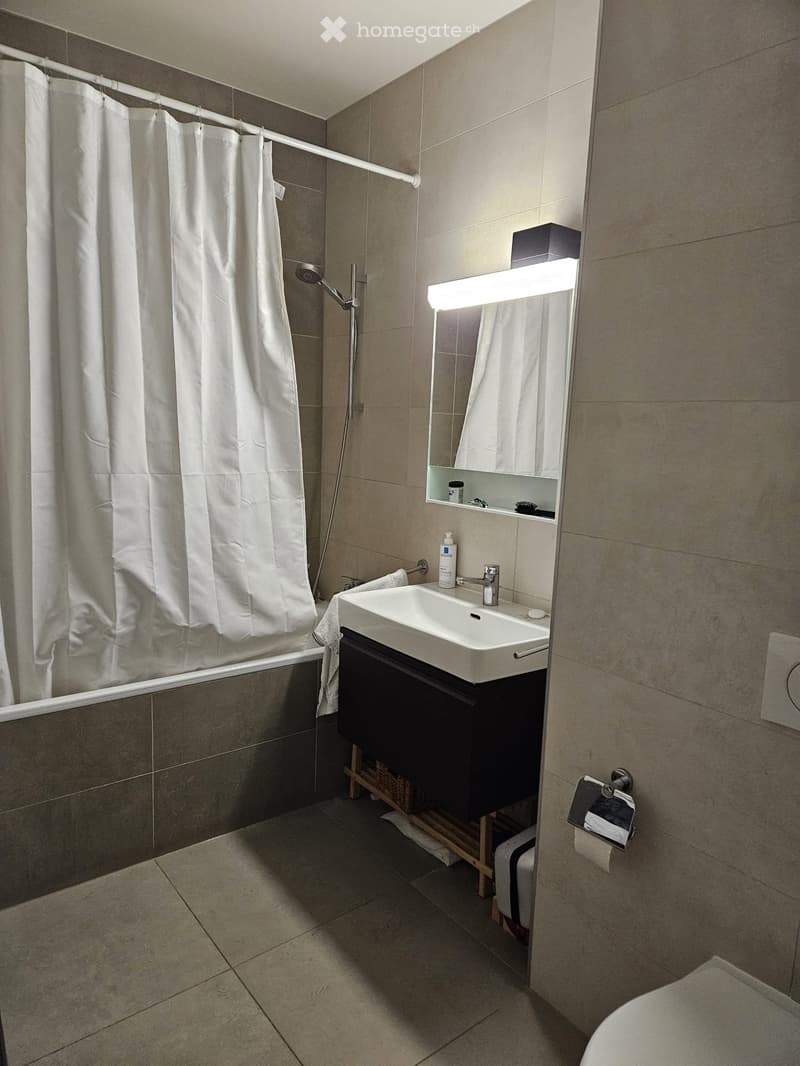 Modern bathroom with a white sink, mirror, shower curtain, and toilet. Features a sleek design with tiled walls and floor.