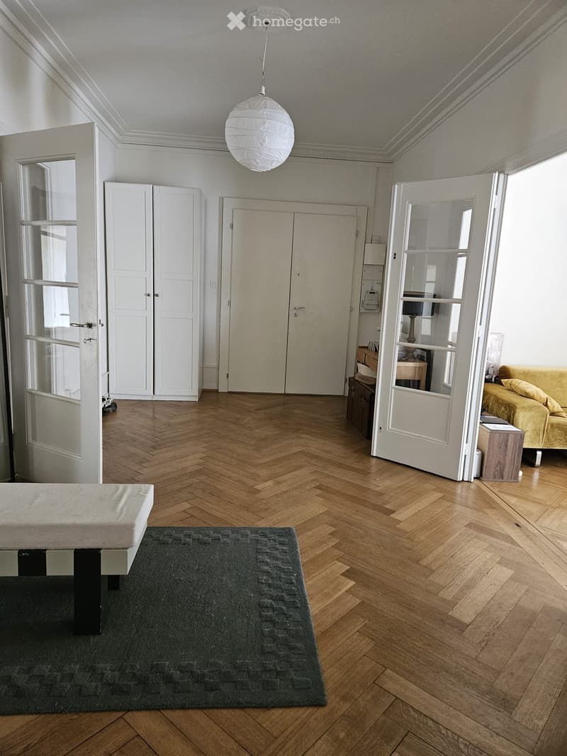 Bright, open room with herringbone wooden flooring, white walls, and large windows. Features a bed, sofa, wardrobe, small table, and a hanging paper lantern light fixture.