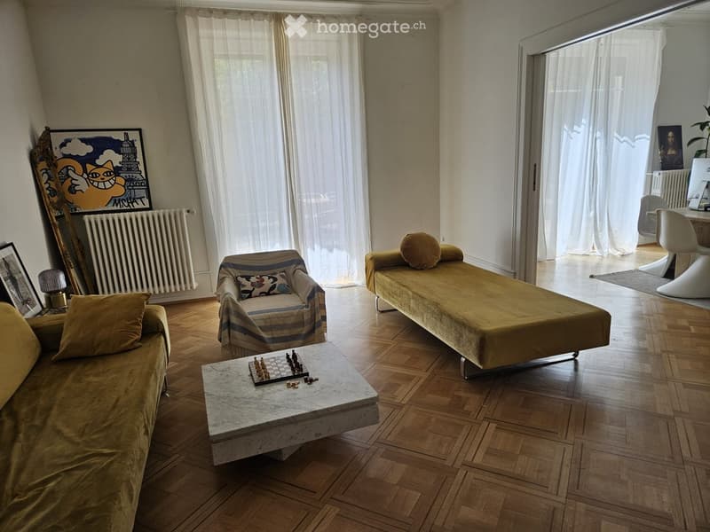 Living room with wooden flooring, large windows with white curtains, a sofa, a chair, a coffee table with a chessboard, and a small table with chairs.