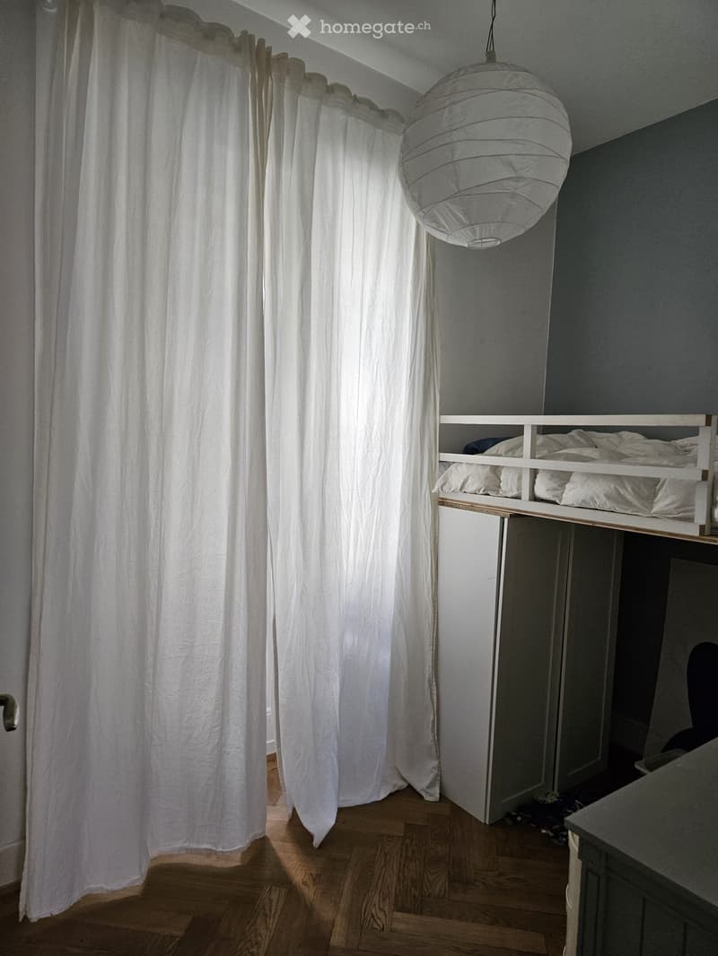 Cozy bedroom with a lofted twin-sized bed, white curtains, and a hanging pendant light. The room features wooden flooring and built-in storage beneath the bed.