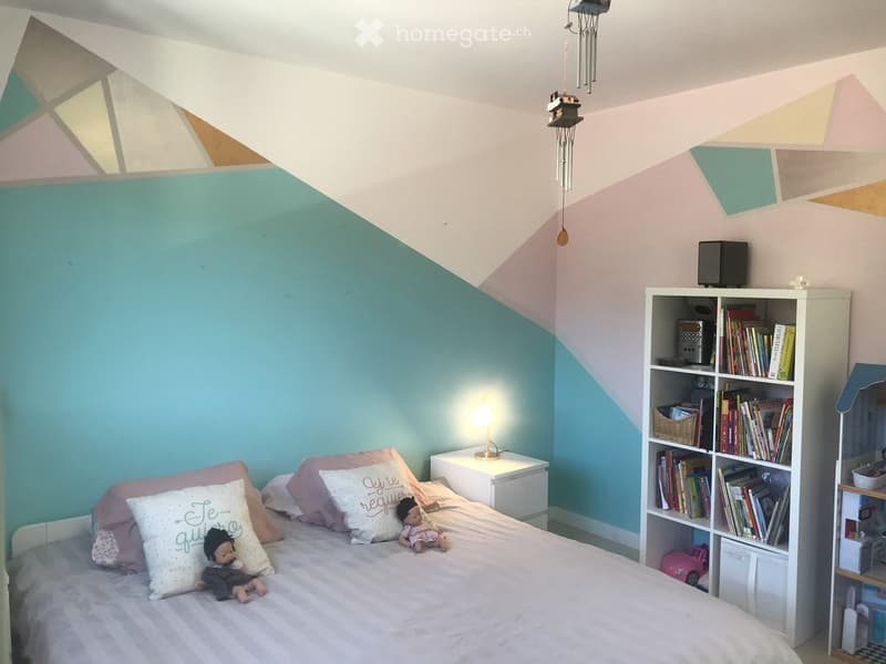 A cozy bedroom with a slanted ceiling, a bed with pillows and a blanket, a nightstand with a lamp, a bookshelf filled with books and a speaker, and decorative items including a wind chime and toys.