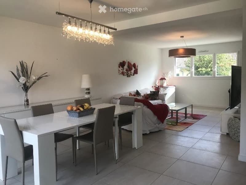 Modern, open-plan living and dining area with white walls, grey tiled flooring, and contemporary furniture including a sofa, dining table with chairs, coffee table, and decorative lamps.