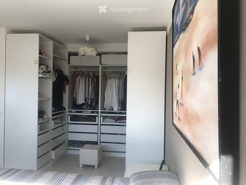 Modern bedroom with a large wardrobe, neatly organized with hanging clothes and drawers, a small white stool, and a large picture frame on the wall.
