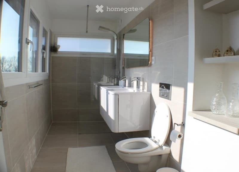 Modern bathroom with a toilet, sink, mirror, and shower area. Features include white cabinetry, a large window, and decorative shelves with vases.