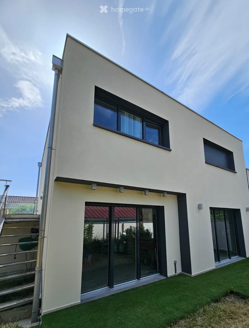 Modern two-story house with light beige exterior, large windows, and a raised ground floor entrance. Features include a staircase leading to the upper level and a small patch of artificial grass in the foreground.
