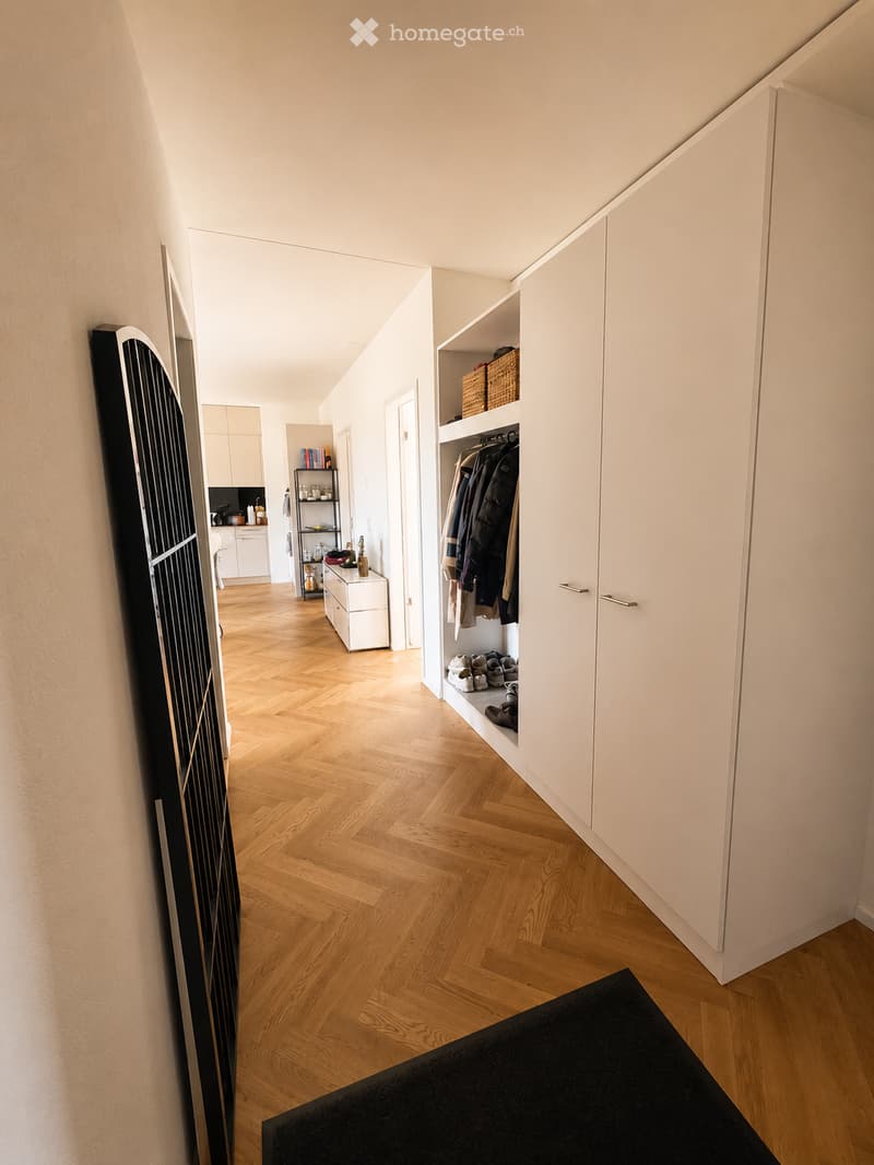 A modern hallway with wooden herringbone flooring, white walls, built-in closet with hanging space, and a small shelf with baskets. The hallway leads to a kitchen area with white cabinets and a countertop.