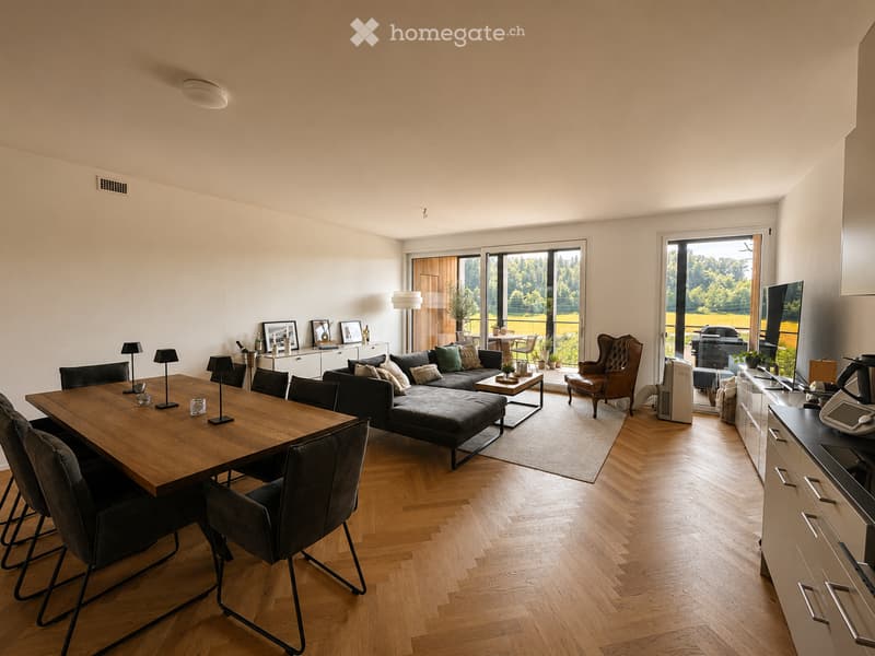 Open-plan living and dining area with wooden flooring, large windows, a sectional sofa, coffee table, dining table with chairs, and a lamp.