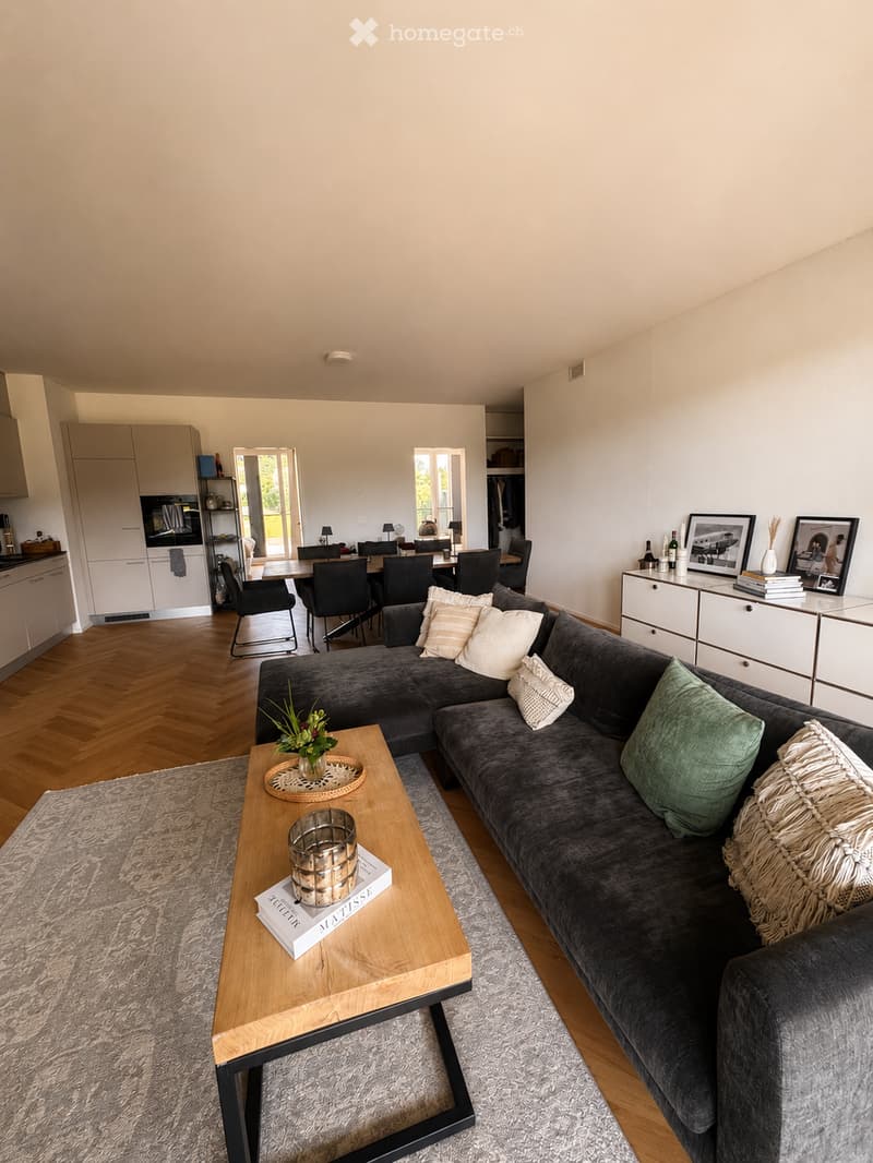 Modern living room with a grey sectional sofa, wooden coffee table, books, vase, and a view of a dining area with chairs and a table. The room features light-colored walls, wooden flooring, and a kitchen area with white cabinets.