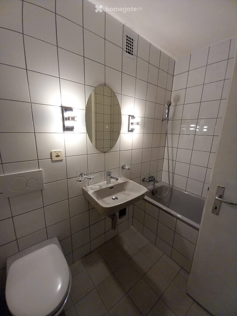 Modern bathroom with white tiled walls and floor, featuring a sink, toilet, bathtub, and showerhead. Includes a mirror and two cups on the sink counter.