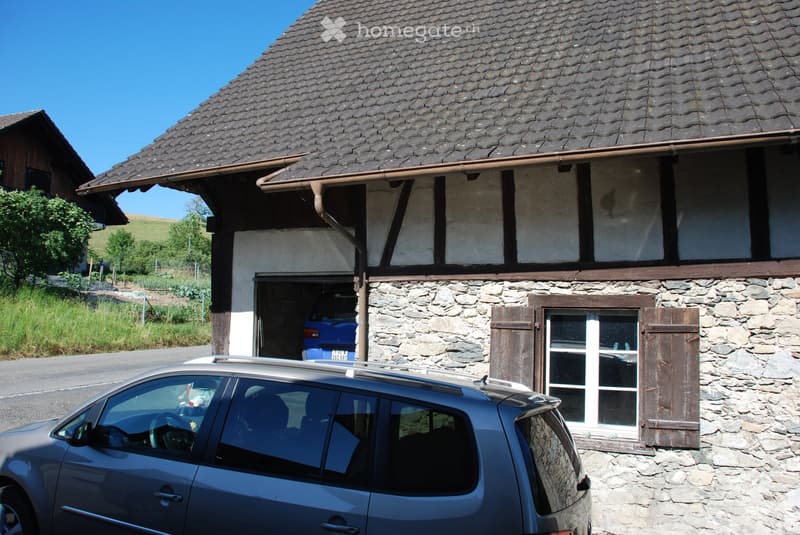 Traditional stone house with a tiled roof, wooden shutters, and a garage. A blue car is parked in the garage, and another car is parked in front of the house.