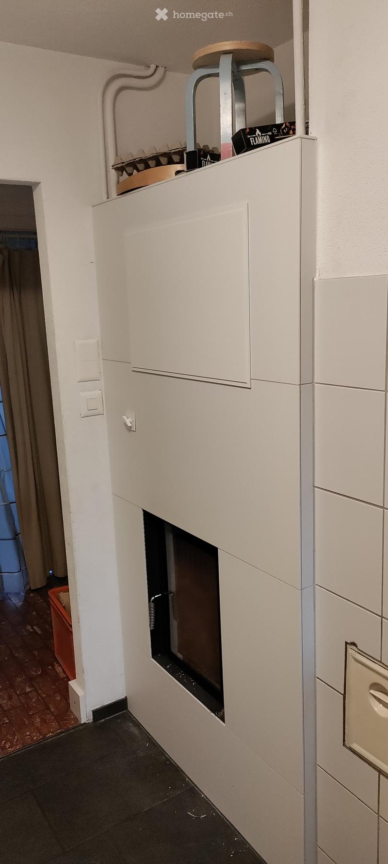 A narrow hallway with white walls, a black tiled floor, and a built-in white cabinet with a small window and a door. There is a light switch on the wall and a small stool on top of the cabinet.