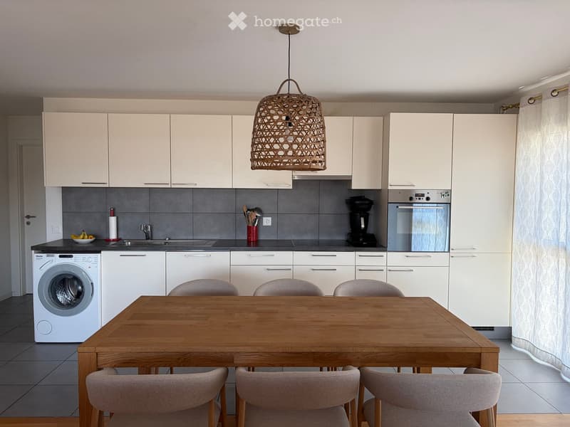 Modern kitchen with white cabinets, grey tiled backsplash, wooden dining table with six chairs, and various appliances including a washing machine, dishwasher, and coffee maker.