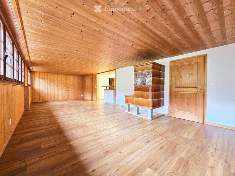 Empty room with wooden floors, wooden walls, and a wooden ceiling. Features a large window, a door, and a small piece of furniture in the background.