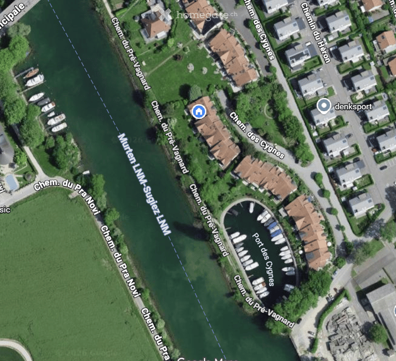 Aerial view of a residential area with houses, a marina, and surrounding greenery. The area includes a waterfront with boats and a network of streets.