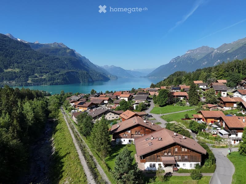 Aerial view of a picturesque village nestled in a valley surrounded by mountains and a lake. Traditional wooden houses with red-tiled roofs are scattered across the landscape, connected by winding roads and pathways.