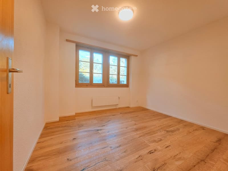 Empty room with wooden flooring, white walls, a large window with wooden frame, a radiator, and a round ceiling light fixture.