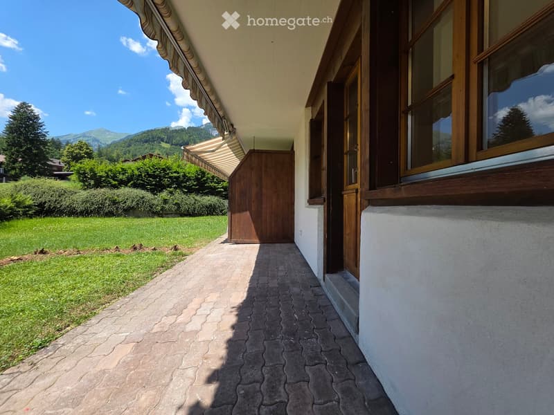 A paved pathway leads to a wooden gate, with a grassy lawn and trees on one side and a house with large windows on the other. The background shows a scenic mountain view.