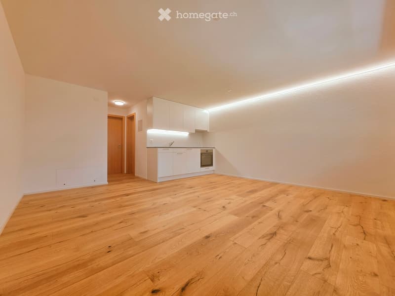 Empty room with light wooden flooring, white walls, and a small white kitchen area with cabinets and an oven. The space is well-lit with recessed lighting and has a door leading to another area.