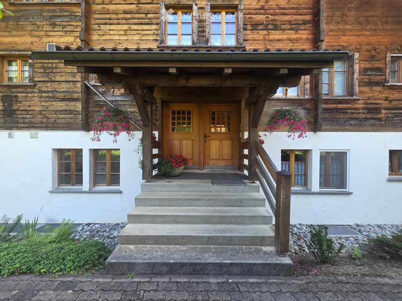 A two-story wooden house with a covered entrance, stone steps, and hanging flower baskets.