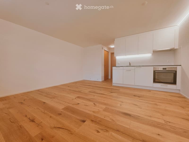 Empty room with light wooden flooring, white walls, and a small kitchen area with white cabinets and a built-in oven.