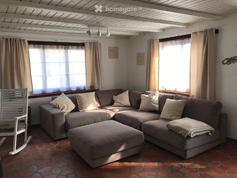 Living room with a large sectional sofa, ottoman, rocking chair, and decorative cushions. The room features exposed wooden beams on the ceiling, large windows with curtains, and a terracotta tile floor.