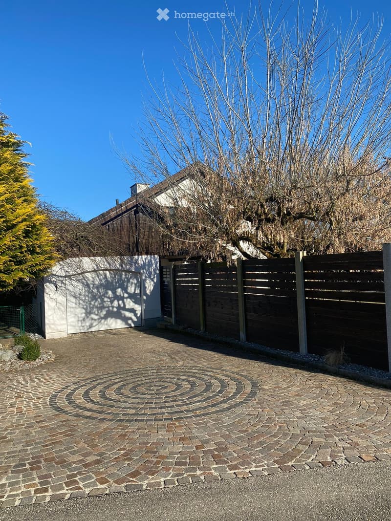 A driveway with a spiral brick pattern leads to a house entrance, surrounded by a wooden fence and bare trees.