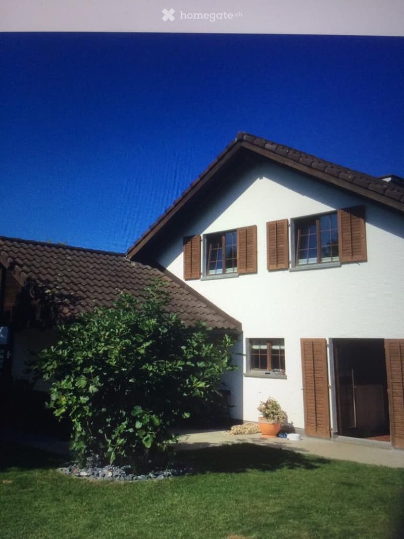 Two-story white house with brown shutters and tiled roof, surrounded by a well-maintained lawn and small garden area.