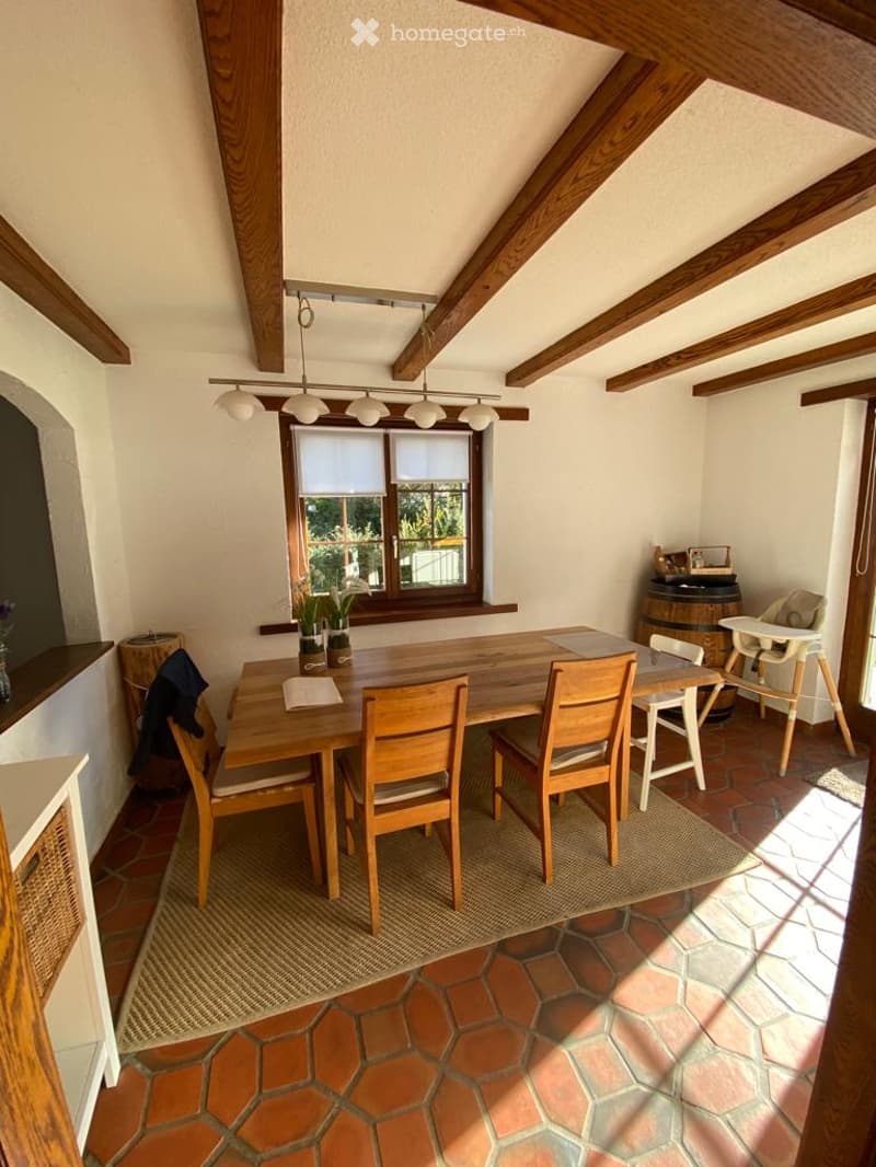 Bright dining area with wooden beams, a large wooden dining table surrounded by chairs, a baby chair, a barrel, and potted plants on the windowsill. The room features exposed wooden beams, a hanging light fixture, and a tiled floor.