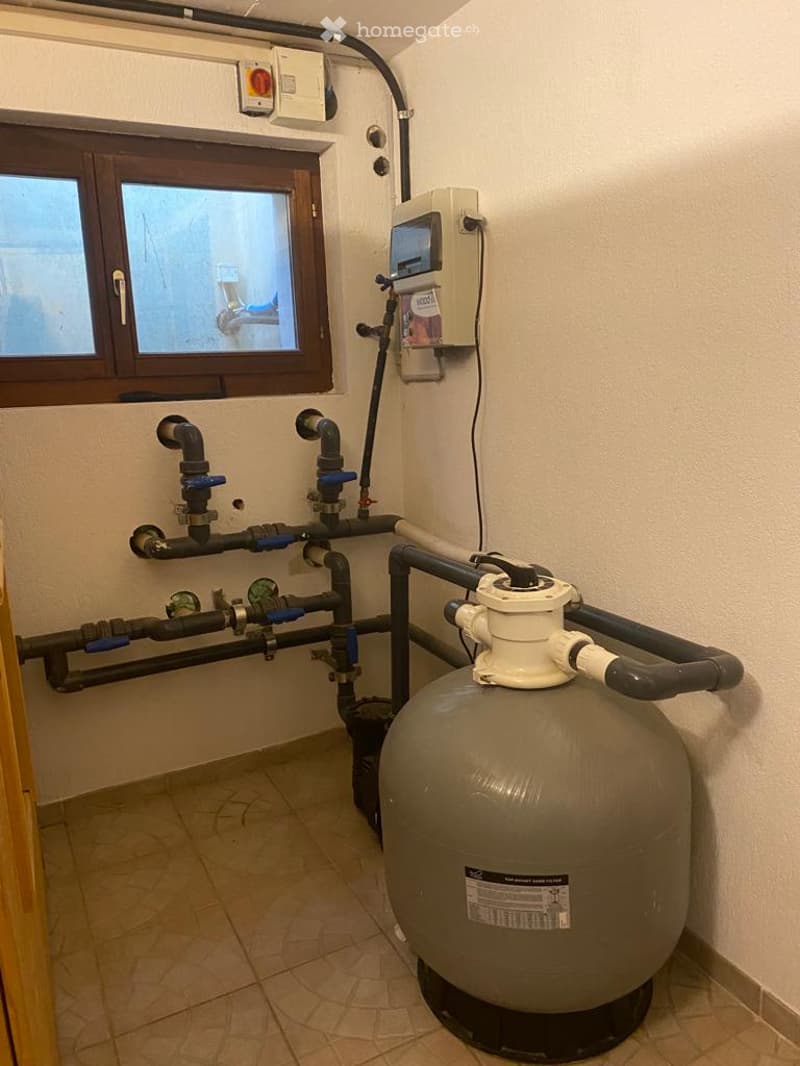 Utility room with water tank, pipes, valves, and control panel. Small window with wooden frame visible.