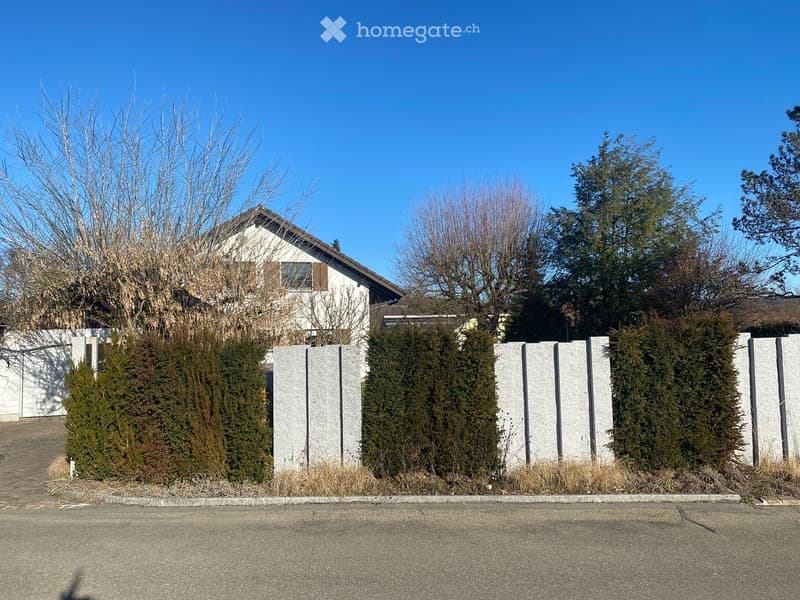 A two-story house with white exterior and brown trim, partially obscured by a stone fence and greenery.