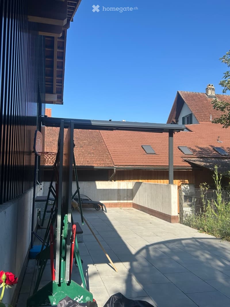 Outdoor patio area with tiled flooring, covered by a pergola, adjacent to a house with a red-tiled roof and solar panels.